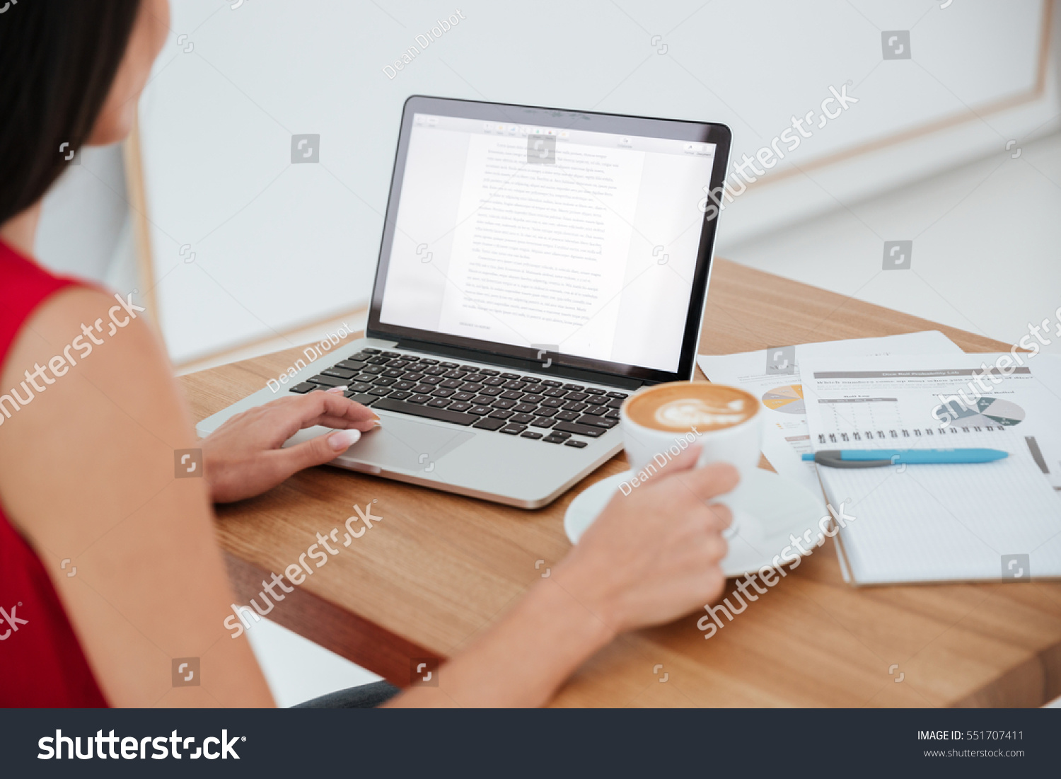 Back view of business woman in red shirt sitting on workplace with laptop, cup of coffee and documents #551707411