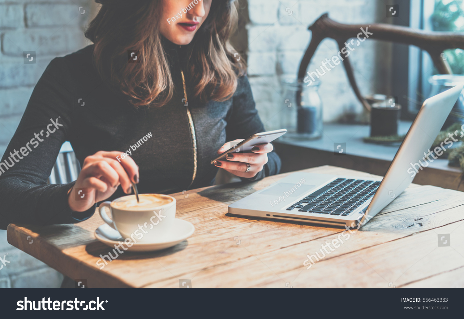 Young woman sitting in coffee shop at wooden table, drinking coffee and using smartphone.On table is laptop. Girl browsing internet, chatting, blogging. Female holding phone and looking on his screen. #556463383