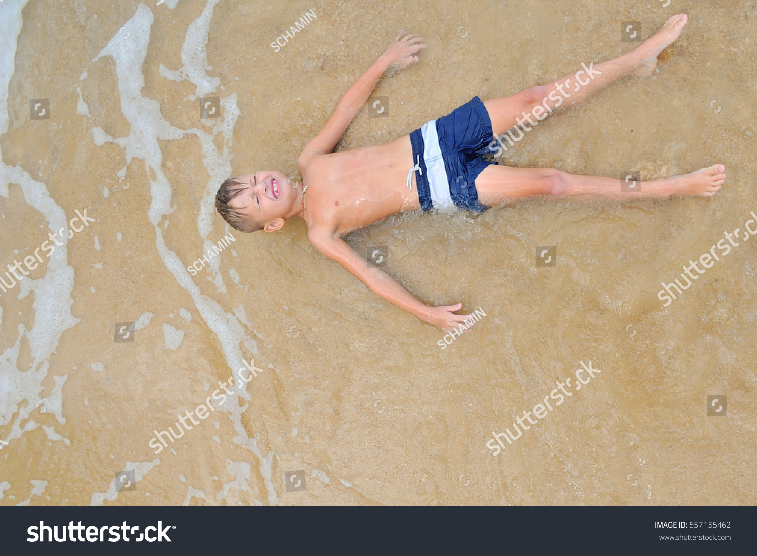 Eight years old handsome blond boy lying on the sand, eyes shut, attracted waves rolls and cover him as if coverlet, view from above #557155462