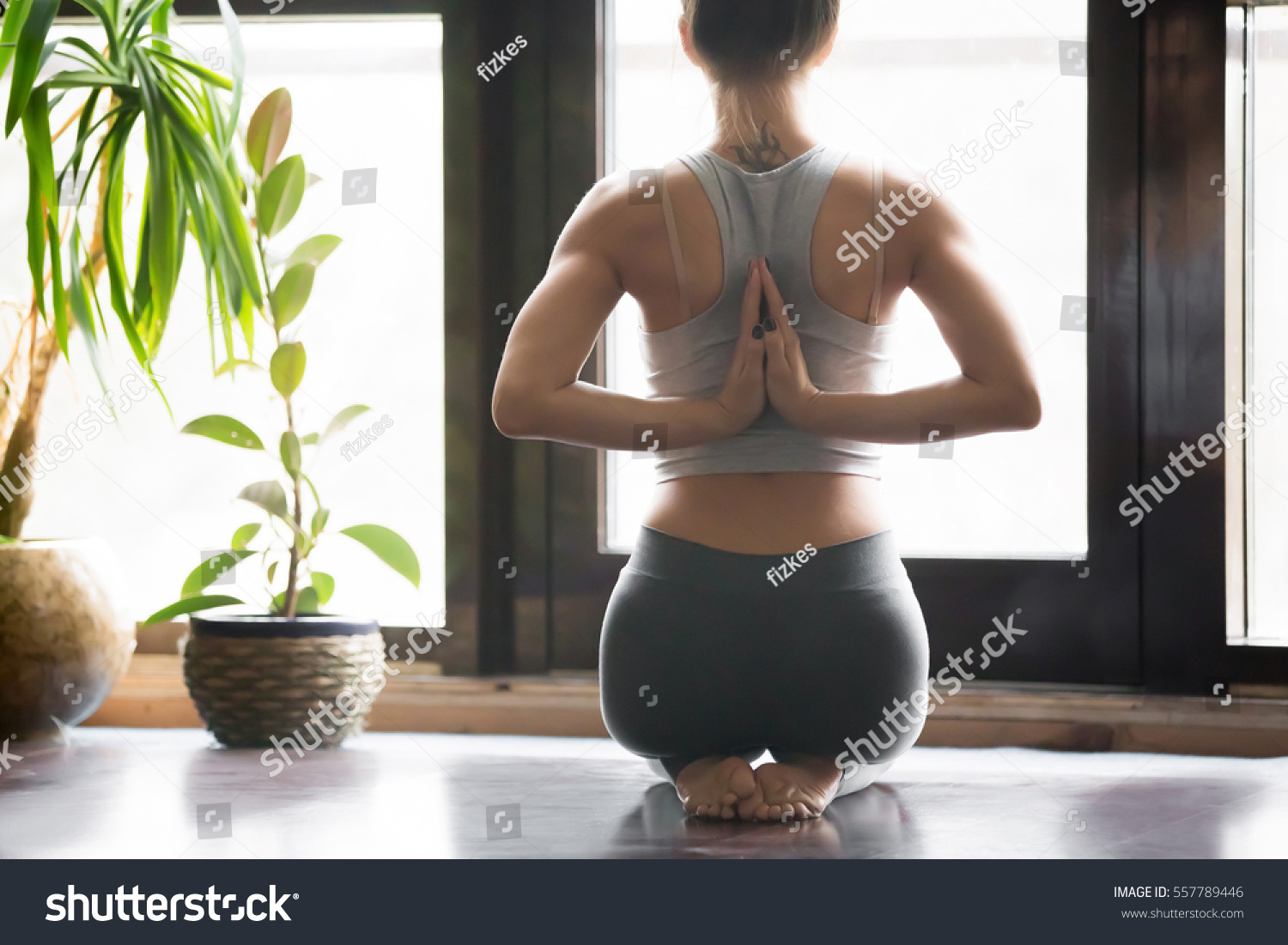 Young woman practicing yoga with namaste behind the back, sitting in seiza exercise, vajrasana pose, working out, wearing sportswear, grey pants, bra, indoor, home interior background, rear view #557789446