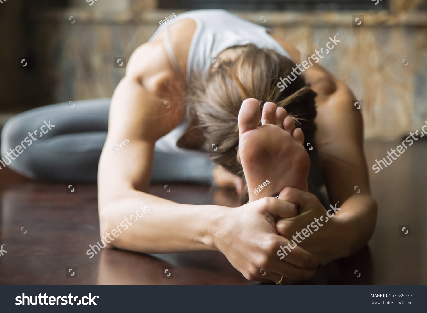 Close up of young woman practicing yoga, sitting in Head to Knee Forward Bend exercise, Janu Sirsasana pose, working out, wearing sportswear, grey pants, bra, indoor, home interior background #557789635