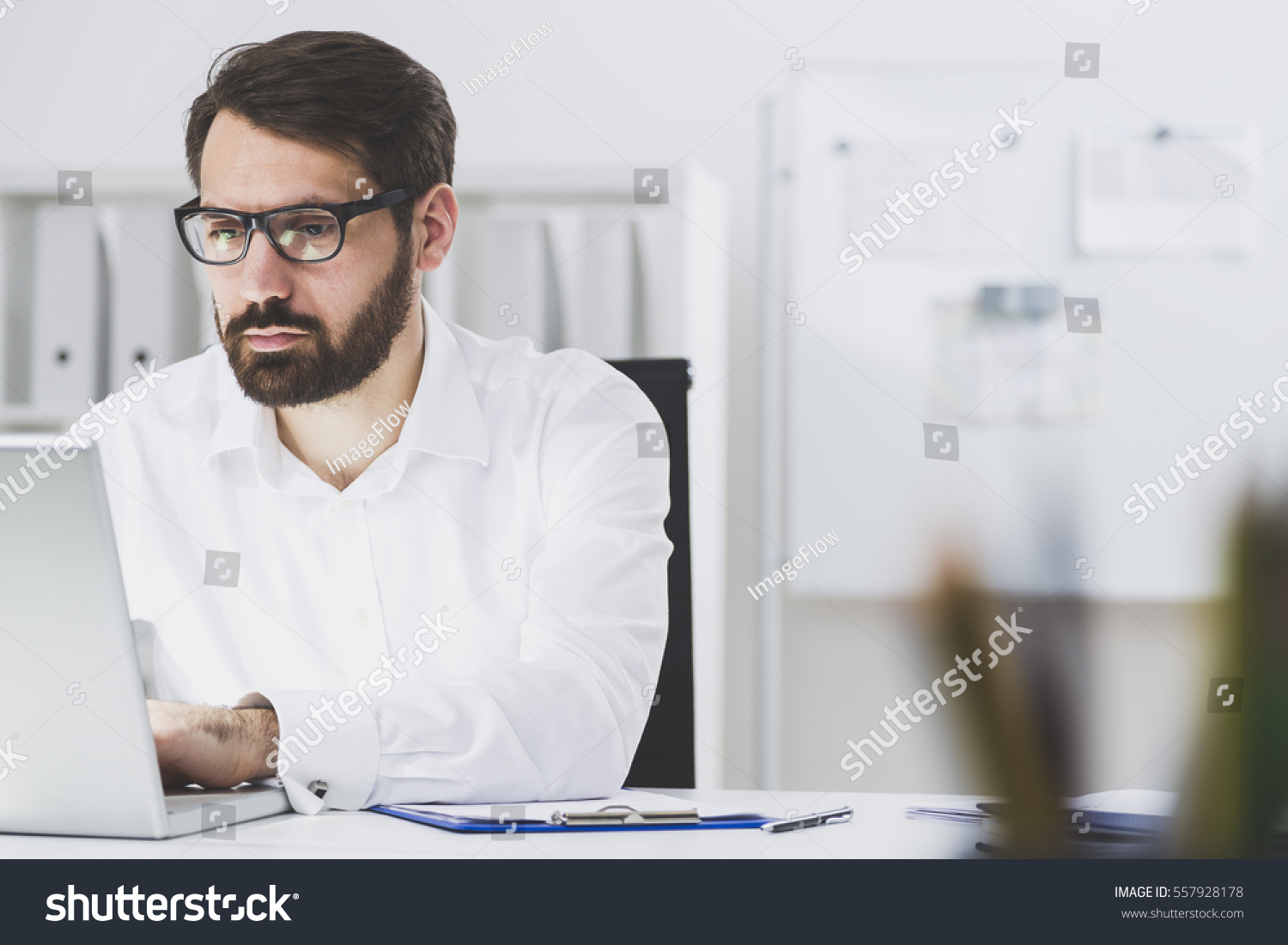 Close up of a bearded businessman wearing glasses and sitting at his laptop and typing. There is a whiteboard and a bookcase in the background. #557928178