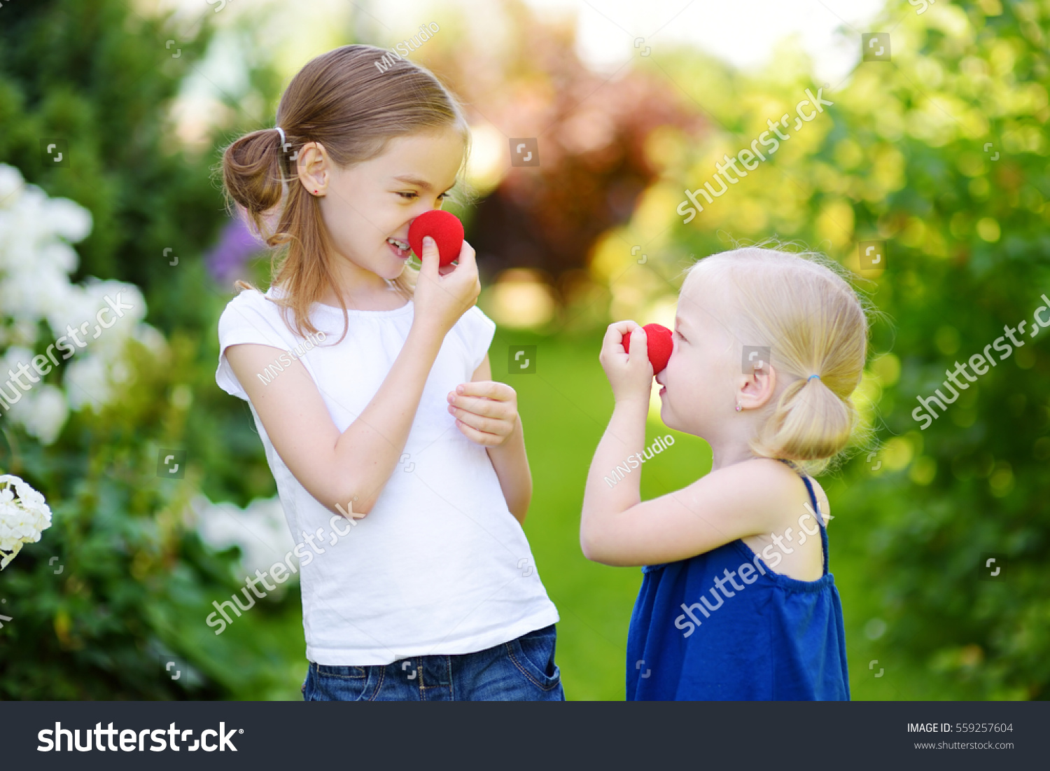 Happy little sisters wearing red clown noses having fun together on sunny summer day outdoors  #559257604