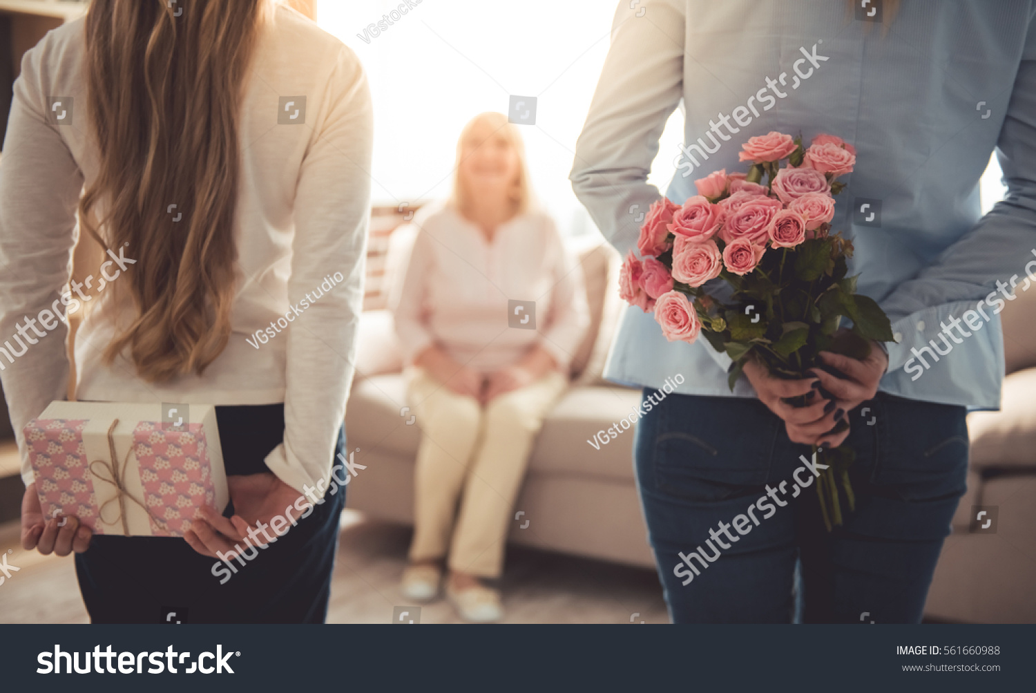 Teenage girl and her mom are hiding flowers and a gift box for their beautiful granny behind backs while grandma is sitting on couch at home #561660988