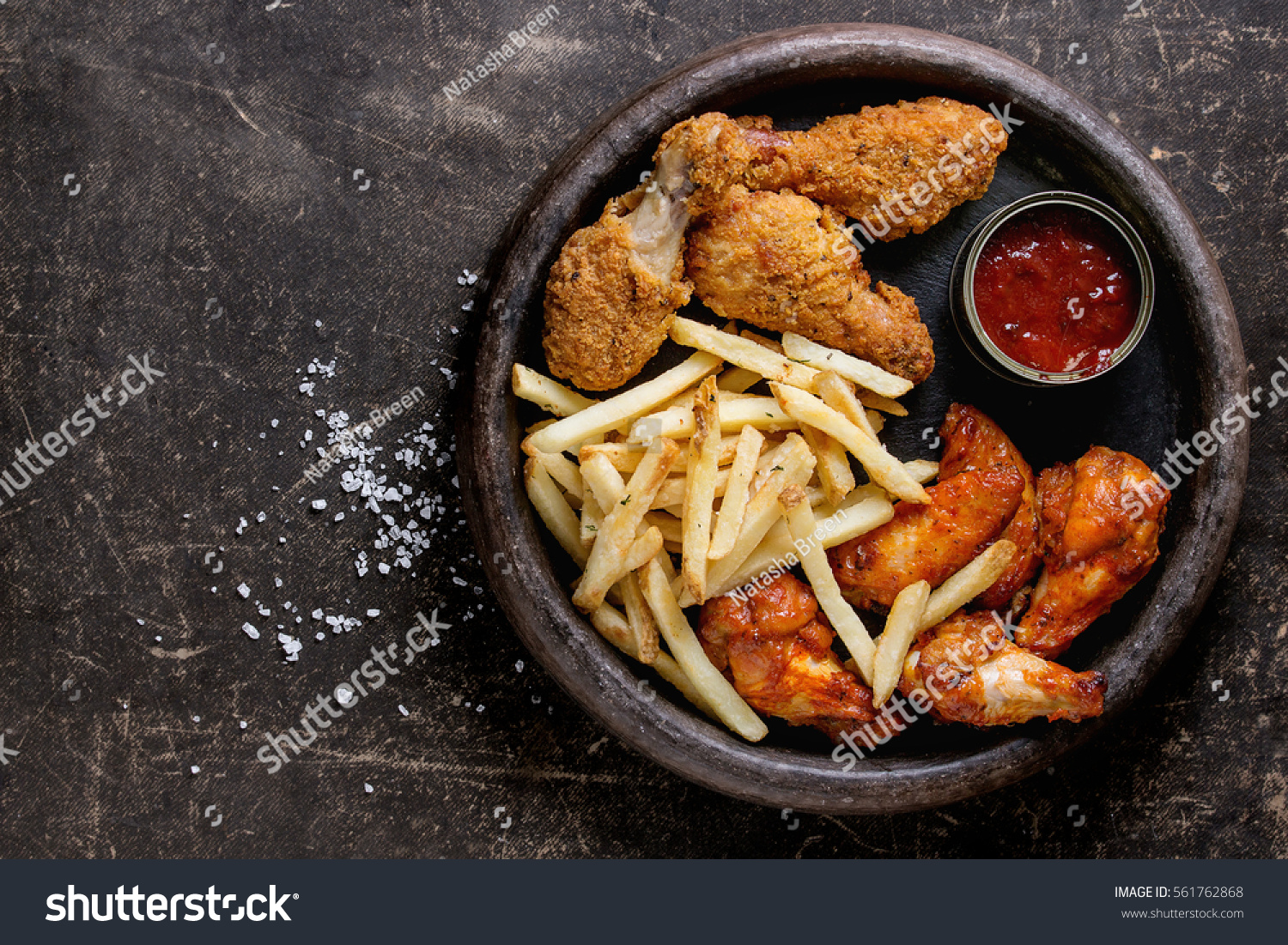 Fast food fried crispy and spicy chicken legs, wings and french fries potatoes with salt and ketchup sauce served in stone plate over dark texture background. Top view, space for text #561762868