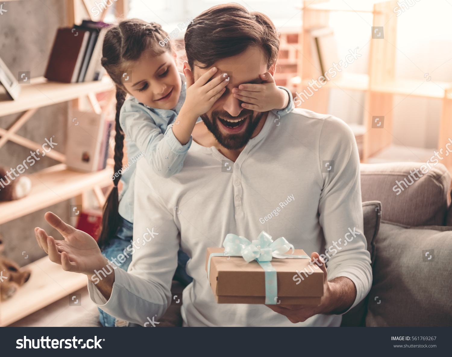 Cute little girl is giving her handsome father a gift box. Both are sitting on couch at home and smiling #561769267
