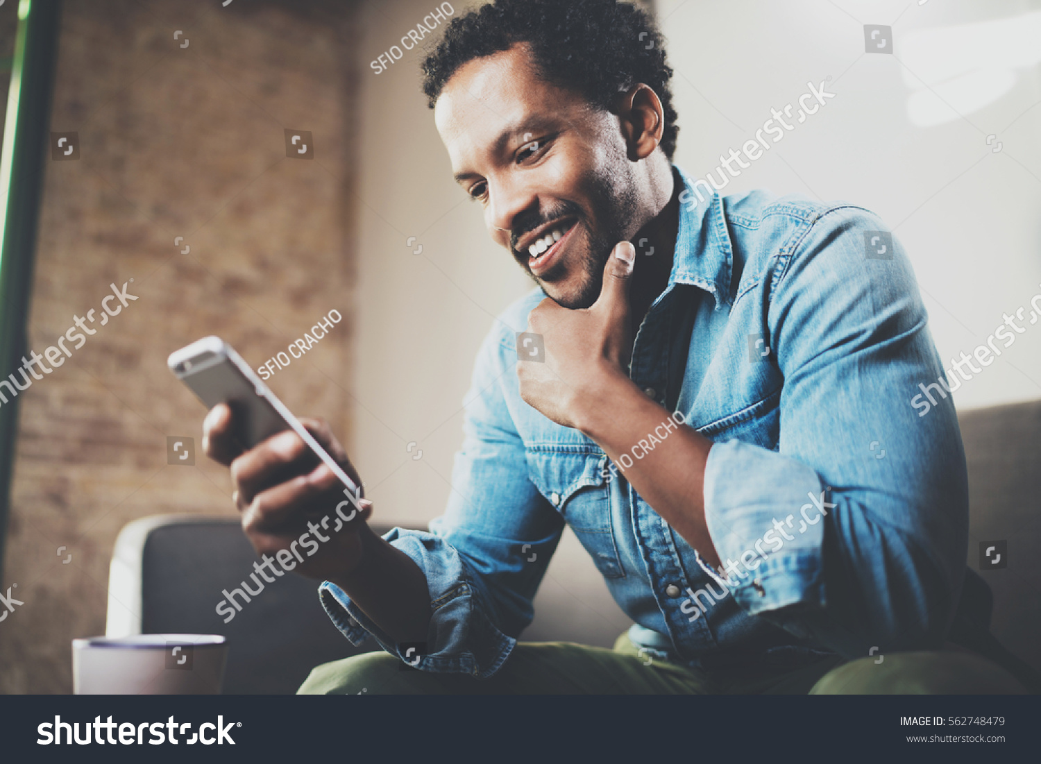 Happy bearded African businessman using phone while sitting on sofa at his modern home.Concept of young people working mobile devices.Blurred background #562748479