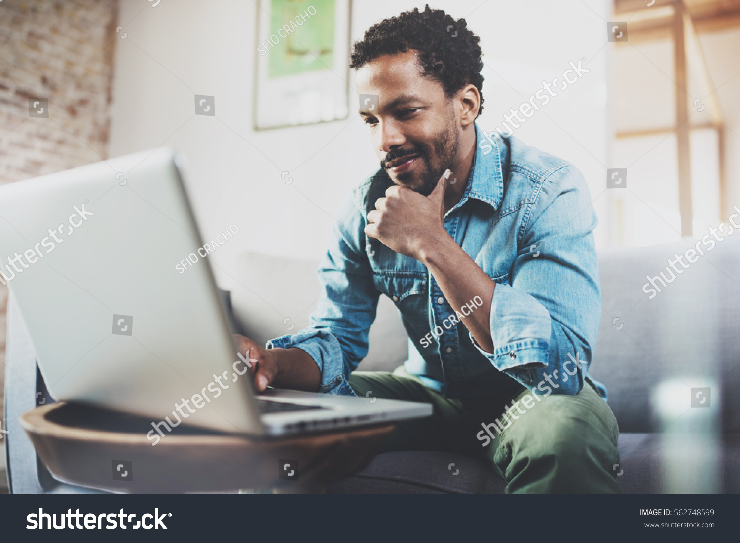 Smiling young African man making video call with friends while sitting on sofa at his modern home.Concept of happy business people.Blurred background, flare effect #562748599