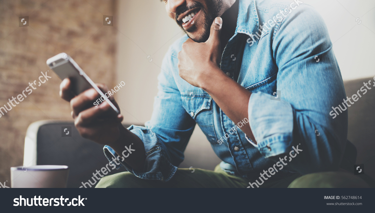Attractive bearded African businessman using smartphone while sitting on sofa at his home.Concept of young people working mobile devices.Closeup with a selected focus.Blurred background.Wide #562748614