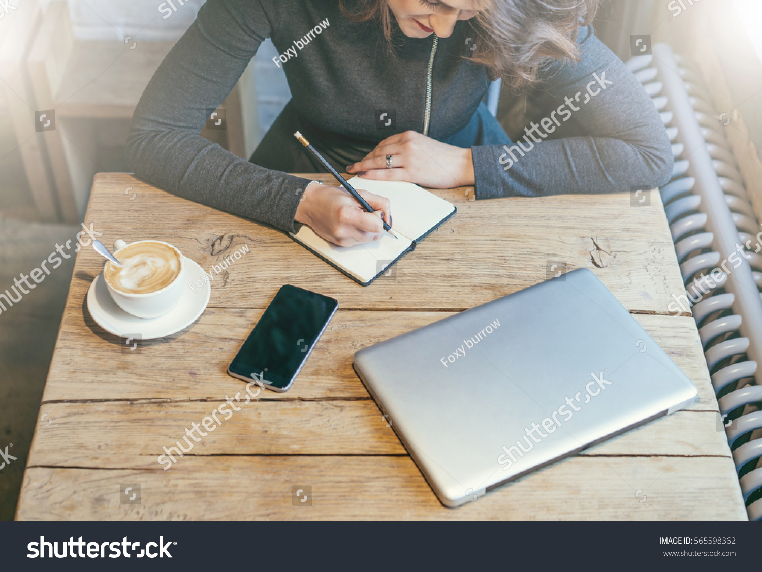 Top view. Young business woman sitting at wooden table in cafe and making notes in notebook. Nearby is cup of coffee, smartphone with blank screen and closed laptop. Student learning online. #565598362