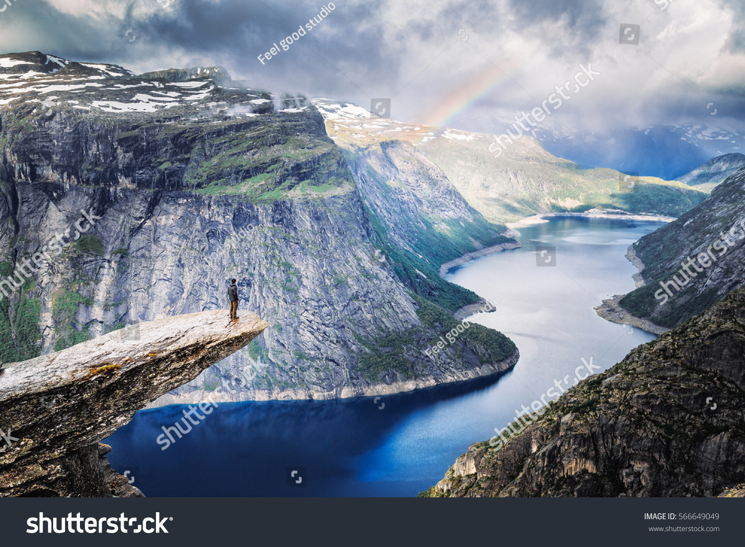Trolltunga (Troll Tongue), climber standing at edge of cliff Trolltunga looking at rainbow against mountains, over amazing blue lake. Beautiful landscape of wild nature in Norway, Scandinavia. #566649049