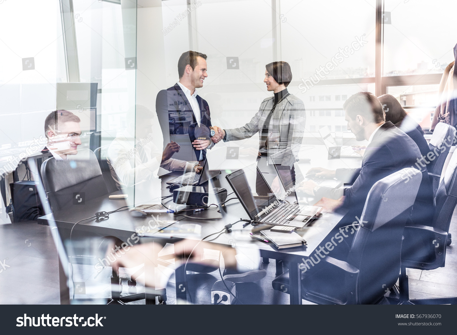 Sealing a deal. Business people shaking hands, finishing up meeting in corporate office. Businessmen working on laptop seen in glass reflection. Business and entrepreneurship concept. #567936070