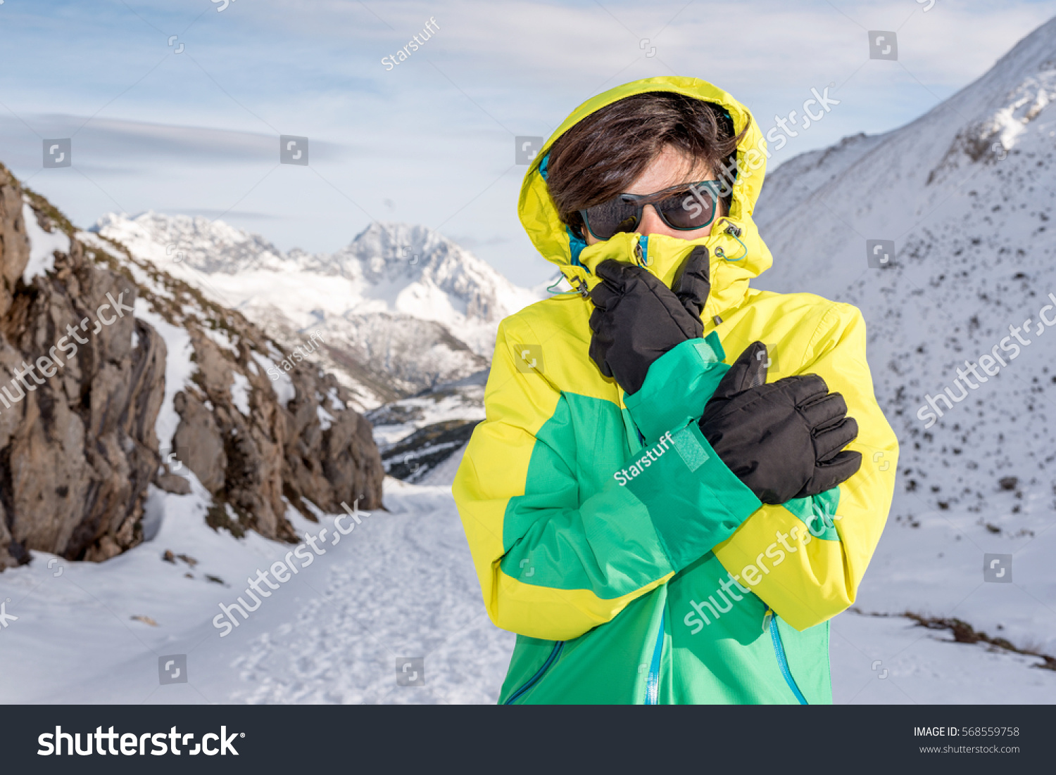 Woman feeling cold discomfort covering her mouth and face from the wind wearing gloves, hood, glasses and windstopper, at extreme snow elements environment on a snowy mountain with low temperature. #568559758