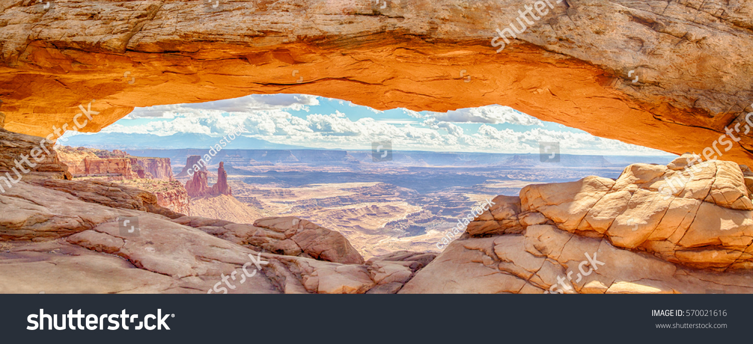 Panoramic view of famous Mesa Arch, iconic symbol of the American West, illuminated golden in beautiful morning light on a sunny day with blue sky and clouds, Canyonlands National Park, Utah, USA #570021616