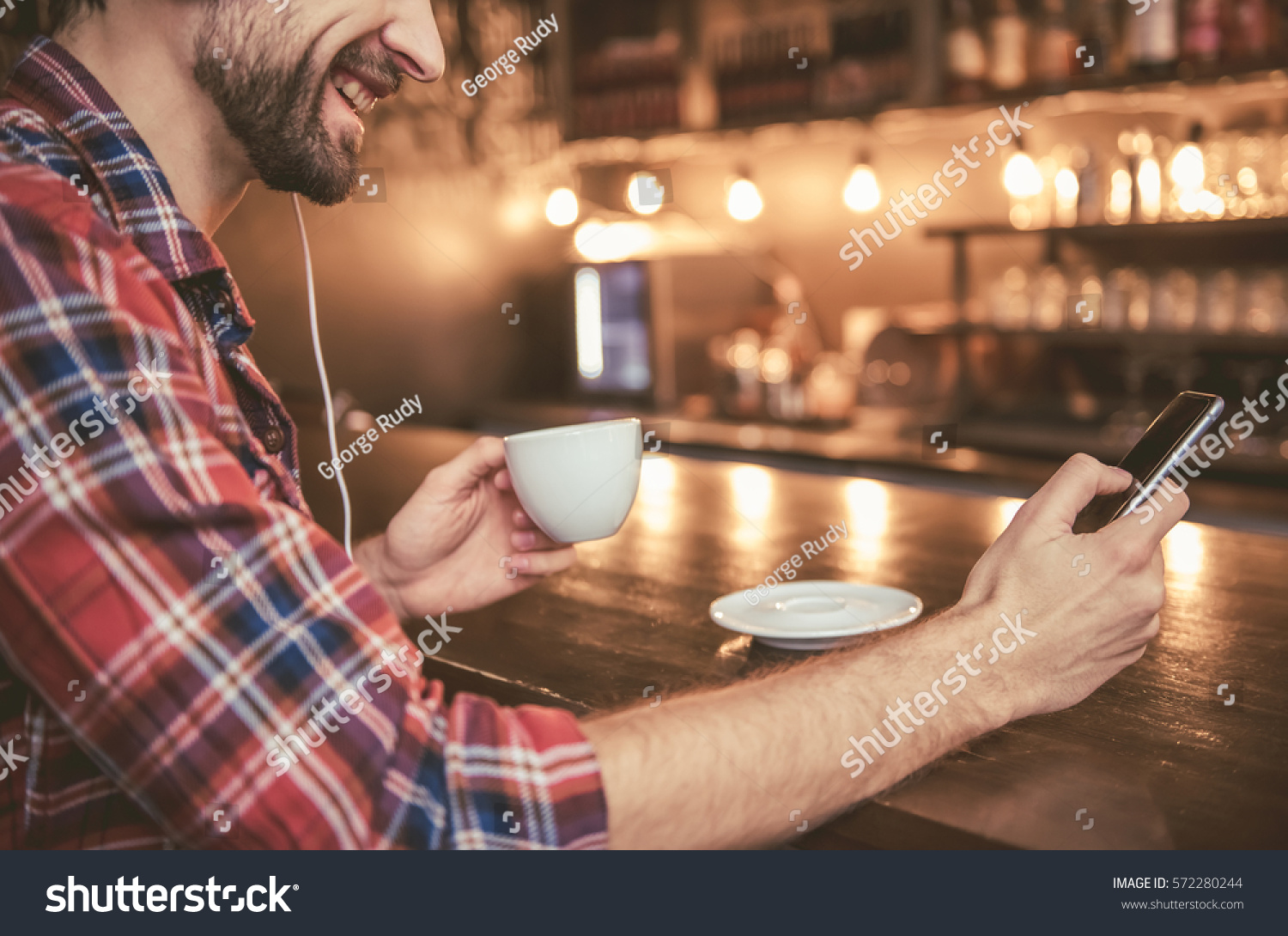Cropped image of man in headphones listening to music using a smartphone, drinking coffee and smiling while resting in cafe #572280244