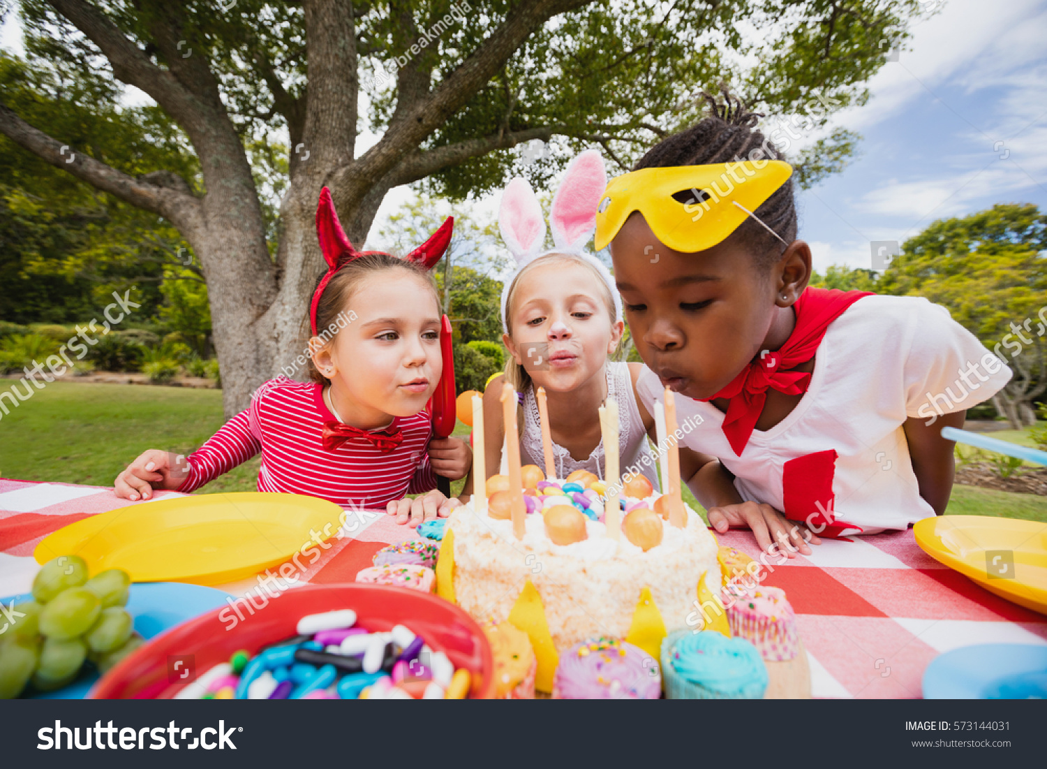 Three little girls blowing together birthday candles in the park #573144031