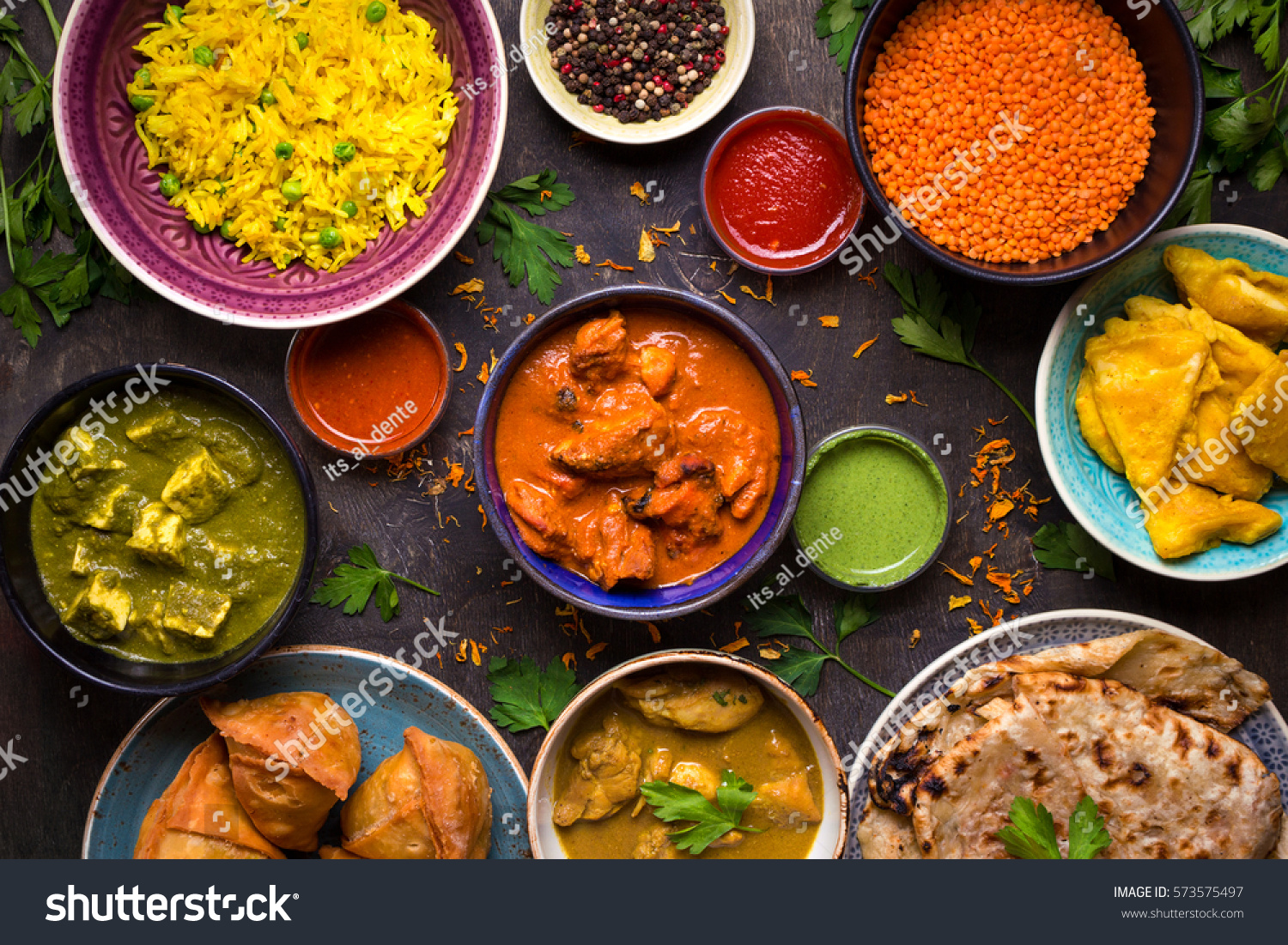 Assorted indian food on dark wooden background. Dishes and appetizers of indian cuisine. Curry, butter chicken, rice, lentils, paneer, samosa, naan, chutney, spices. Bowls and plates with indian food #573575497