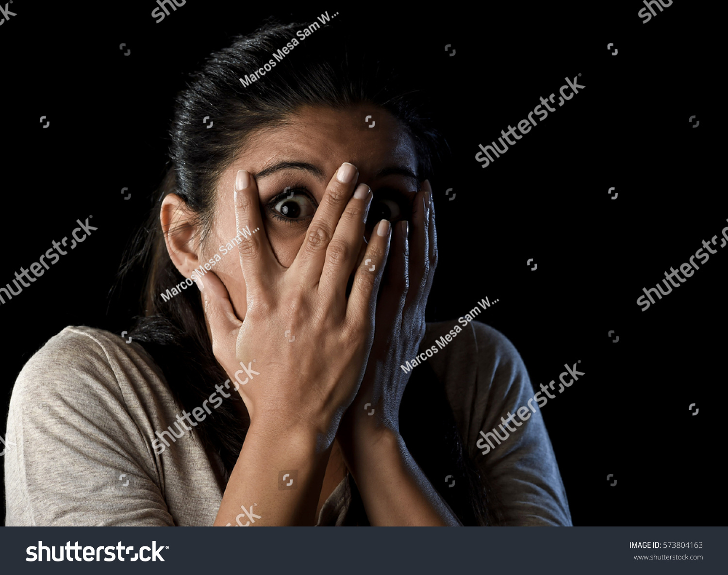 close up portrait young attractive Latin woman desperate and scared isolated on black background looking terrorized and horrified covering her eyes in primal fear emotion face expression #573804163