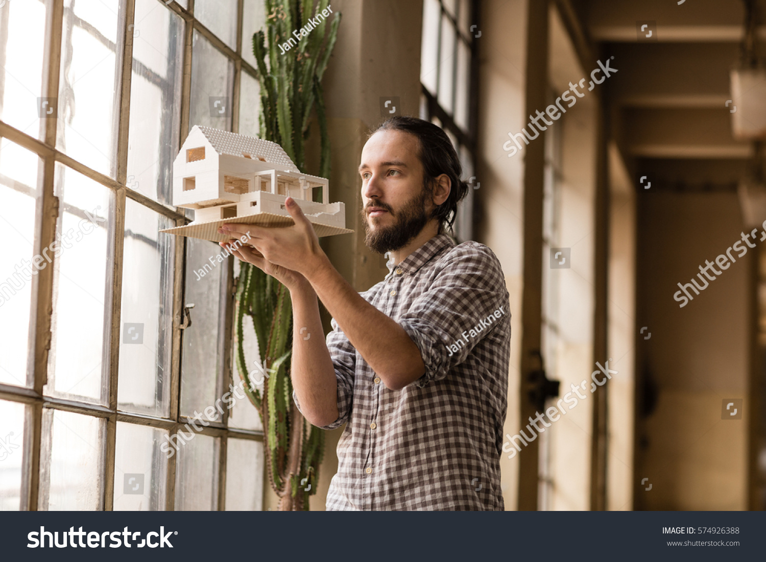 Young architect is looking on the new model in the old industrial space with big factory windows. Man is standing in front of window and holding small model of the house. Color toned image. #574926388