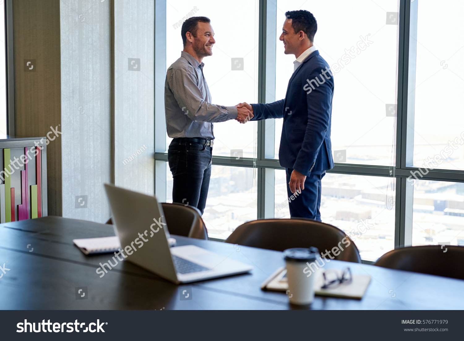 Two smiling businessmen shaking hands together while standing by windows in an office boardroom overlooking the city  #576771979