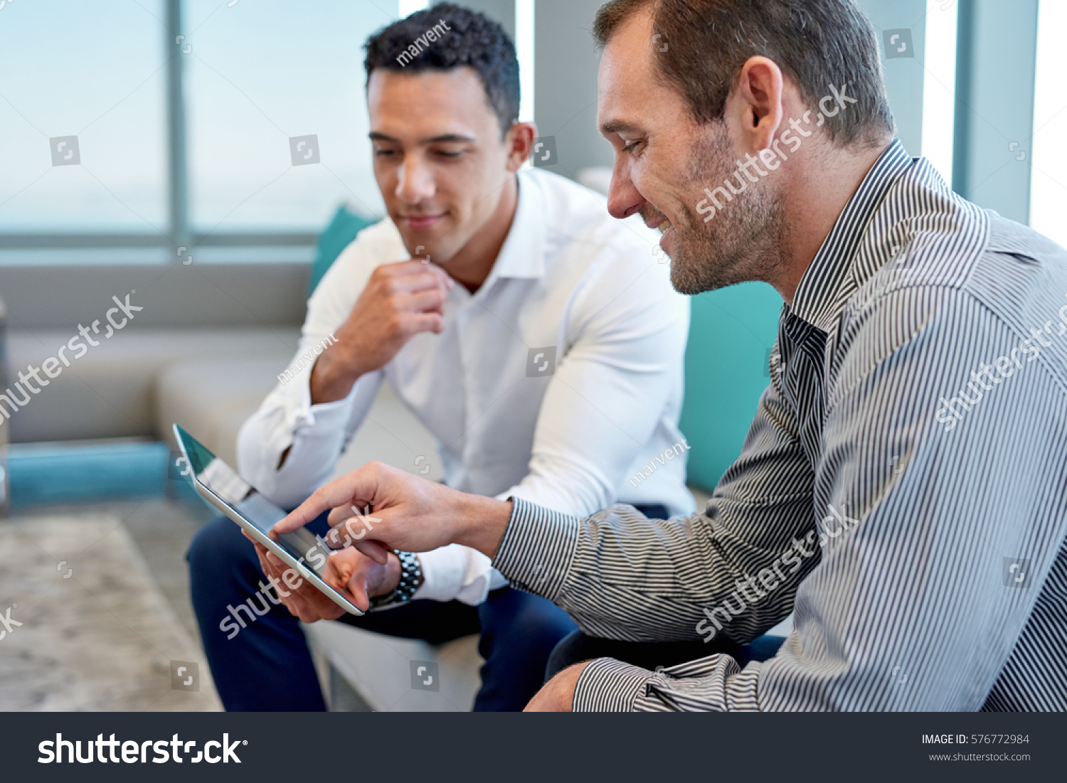 Two smiling male work coworkers sitting on a sofa in a modern office talking together over a digital tablet #576772984