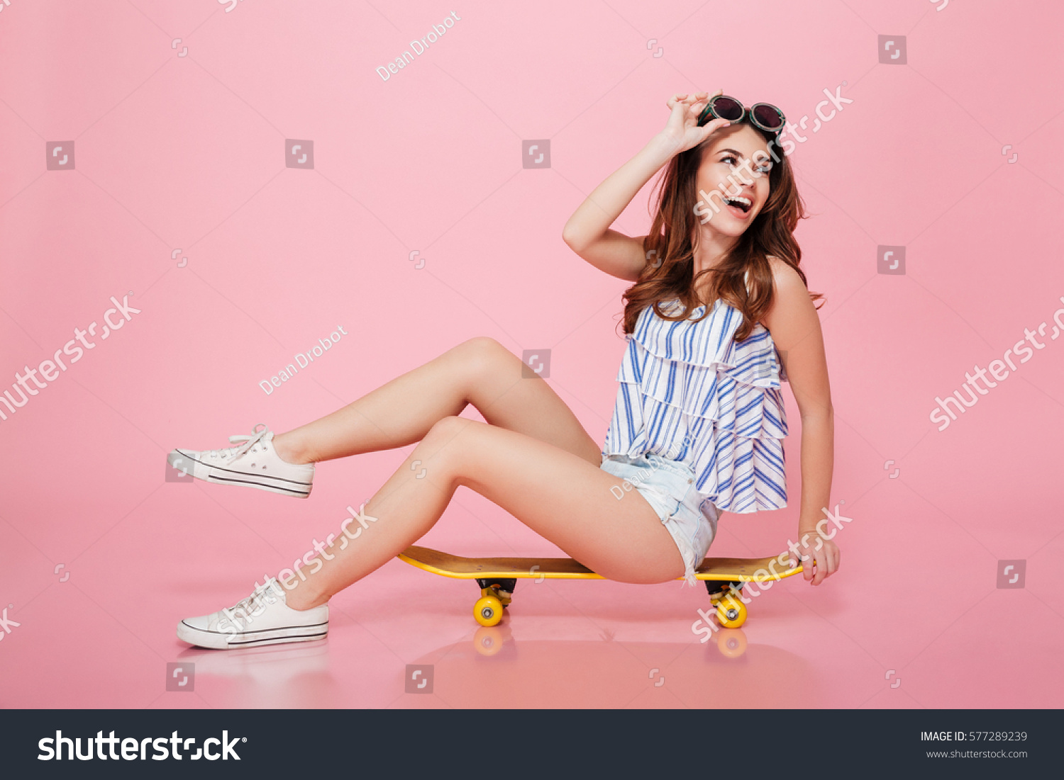 Happy attractive young woman in sunglasses sitting on skateboard over pink background #577289239
