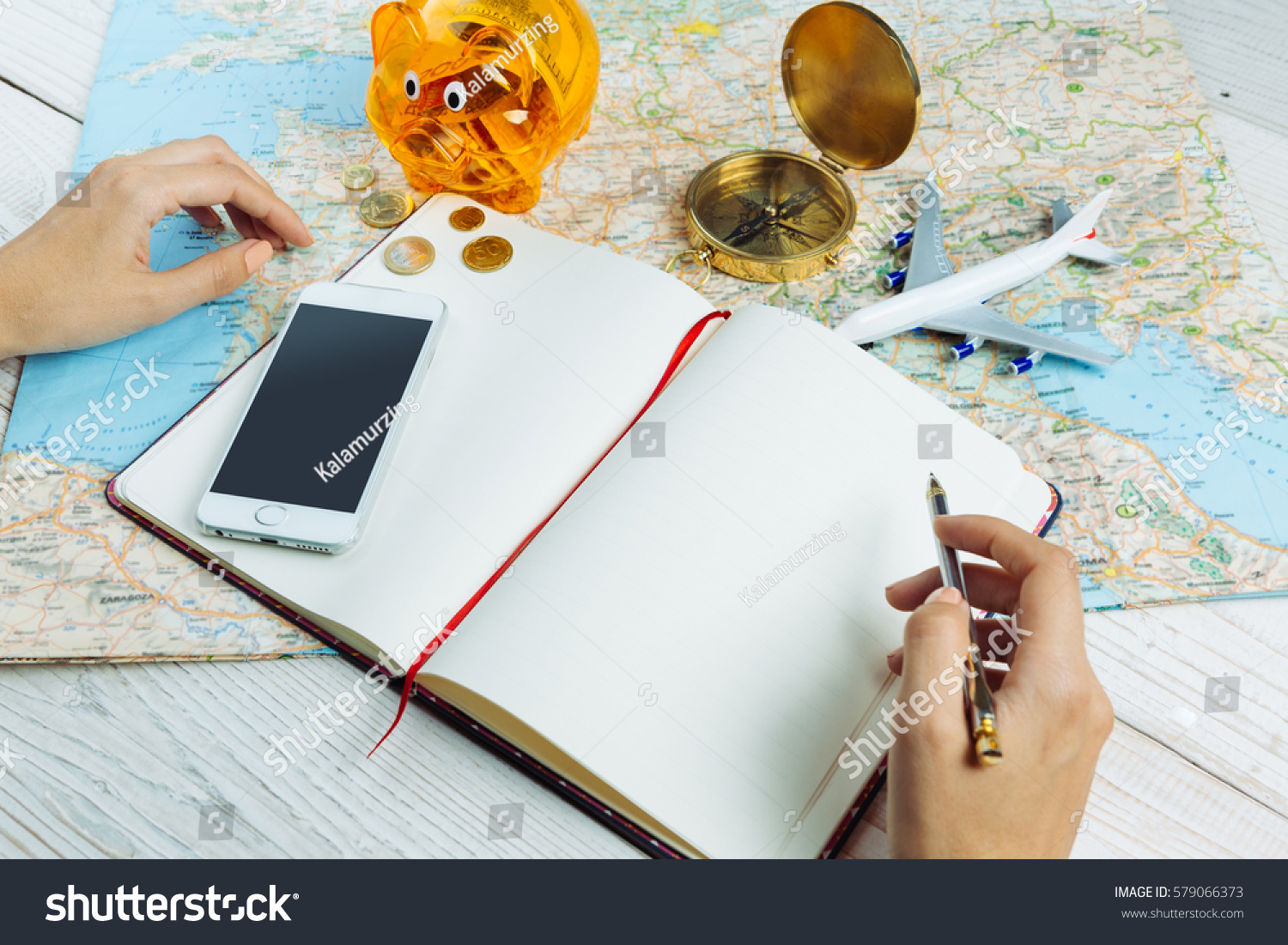Bloger planning budget of trip Hands holding pen near notebook Table with traveling theme items mobile phone, map, compass, plane and piggy bank Empty mockup notes, your can place text logo images.  #579066373