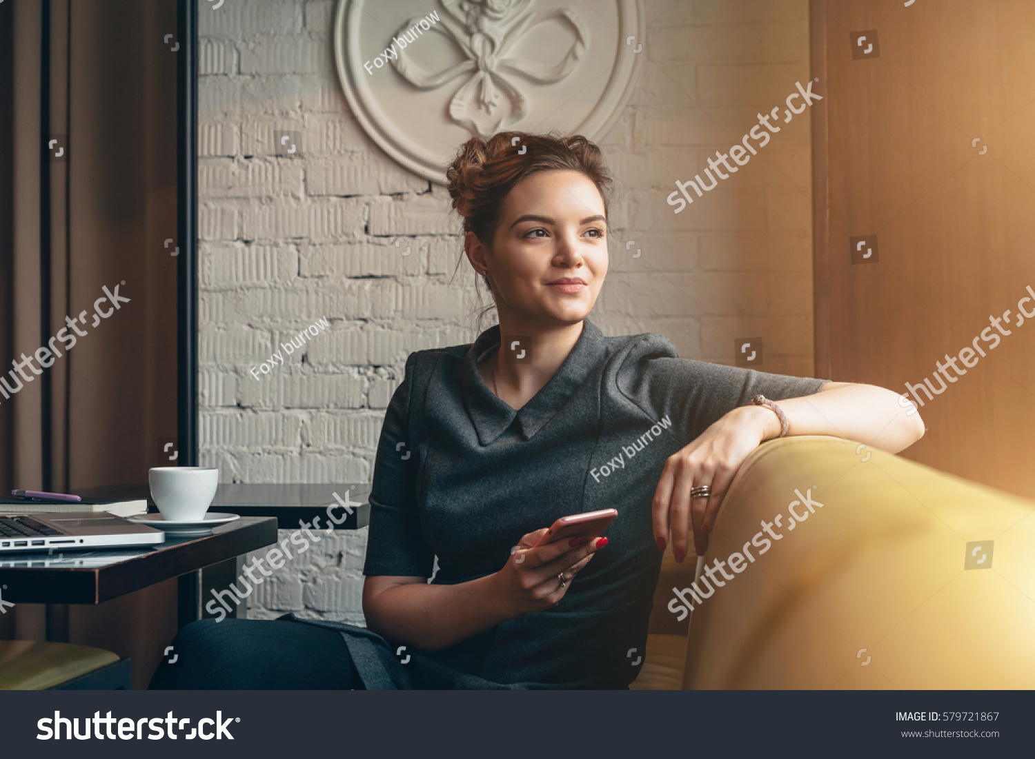 Young smiling business woman sitting in cafe at table, leaning his hand back in chair,looking out window and holding smartphone.On table is laptop, notebook and cup of coffee. Girl using gadget. #579721867