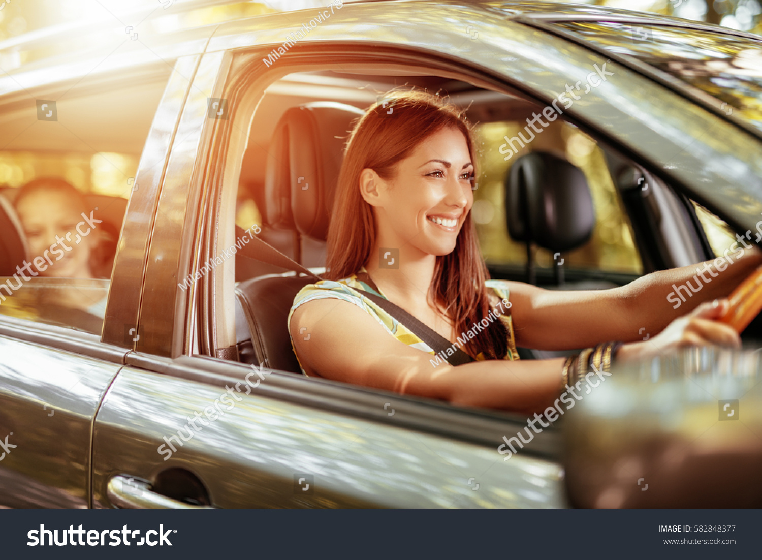 Young beautiful smiling woman driving a car.  Her cute daughter sitting on rear and enjoying. #582848377