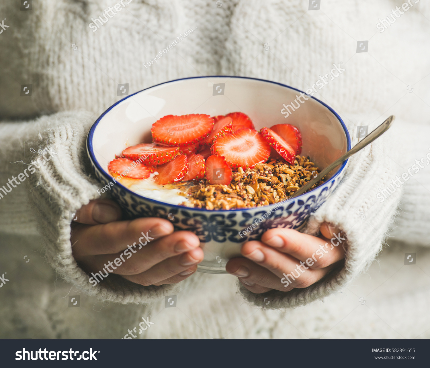 Healthy breakfast greek yogurt, granola and strawberry bowl in hands of woman wearing white loose woolen sweater, selective focus. Clean eating, healthy, vegetarian, dieting food concept #582891655