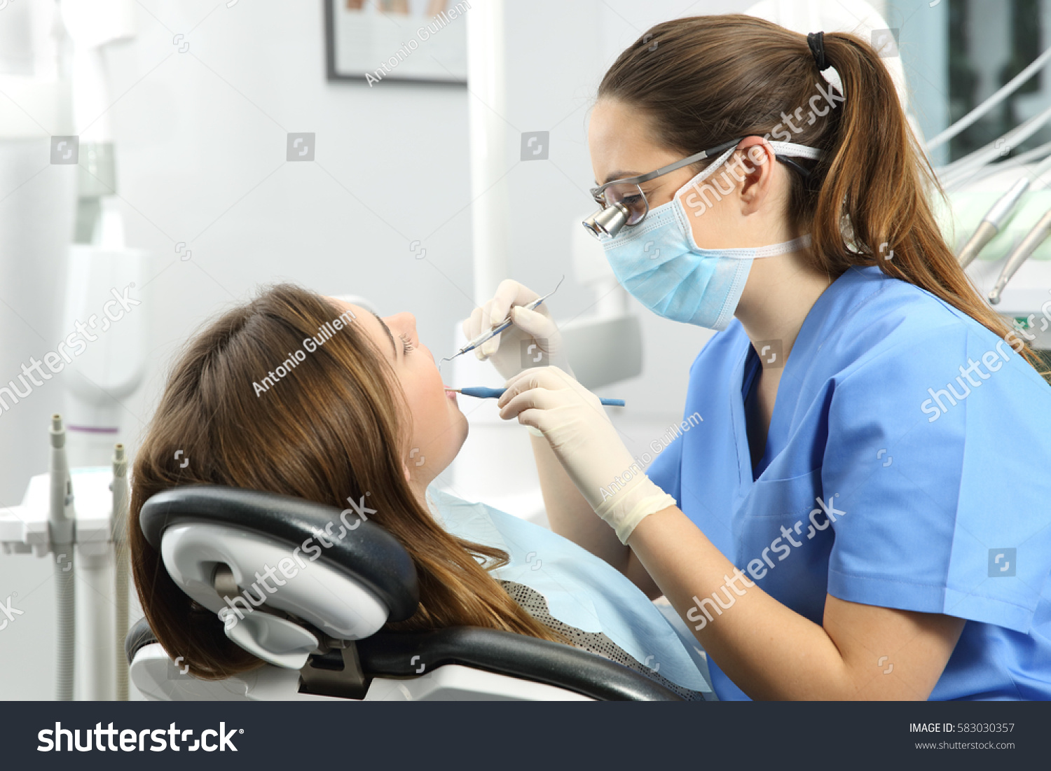 Dentist wearing eyeglasses gloves and mask examining a patient teeth with a dental probe and a mirror in a clinic box with equipment in the background #583030357