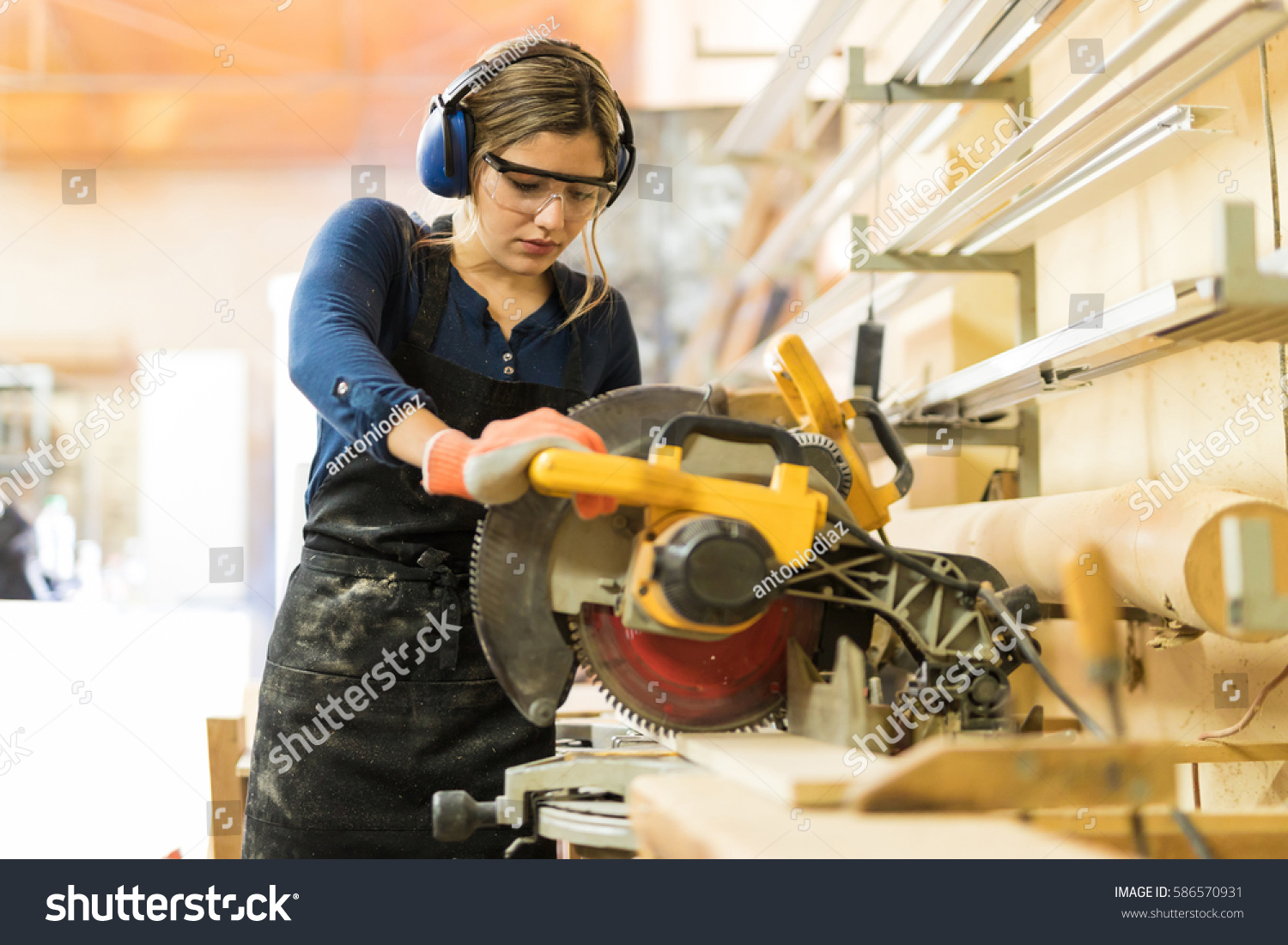 Attractive female carpenter using some power tools for her work in a woodshop #586570931