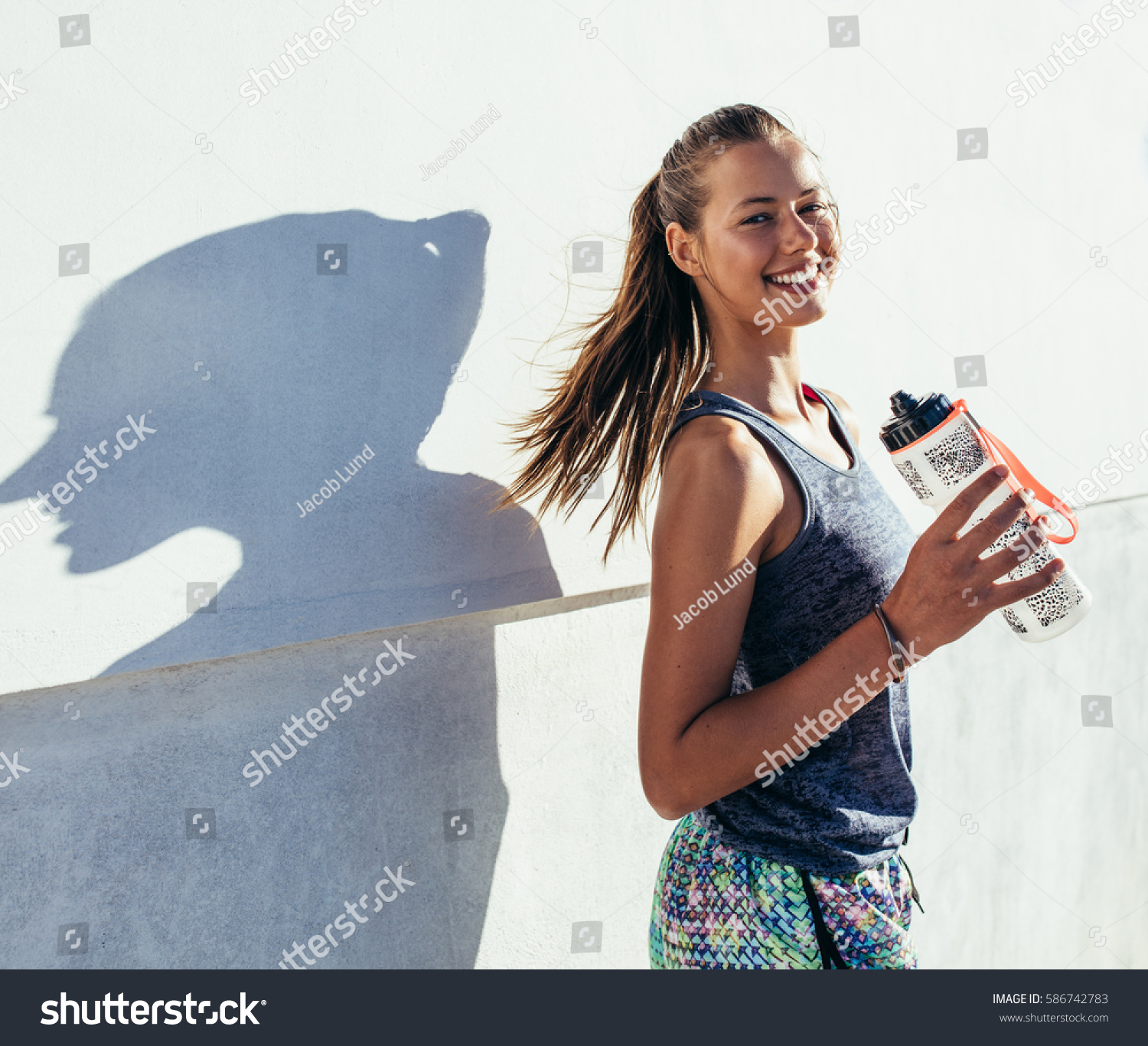 Shot of beautiful female runner standing outdoors holding water bottle. Fitness woman taking a break after running workout. #586742783