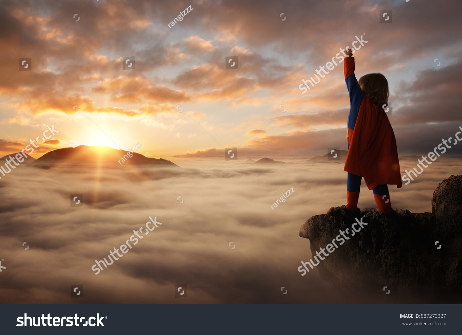 Little boy acting like a superhero on top of the mountain at sunset with copy space #587273327
