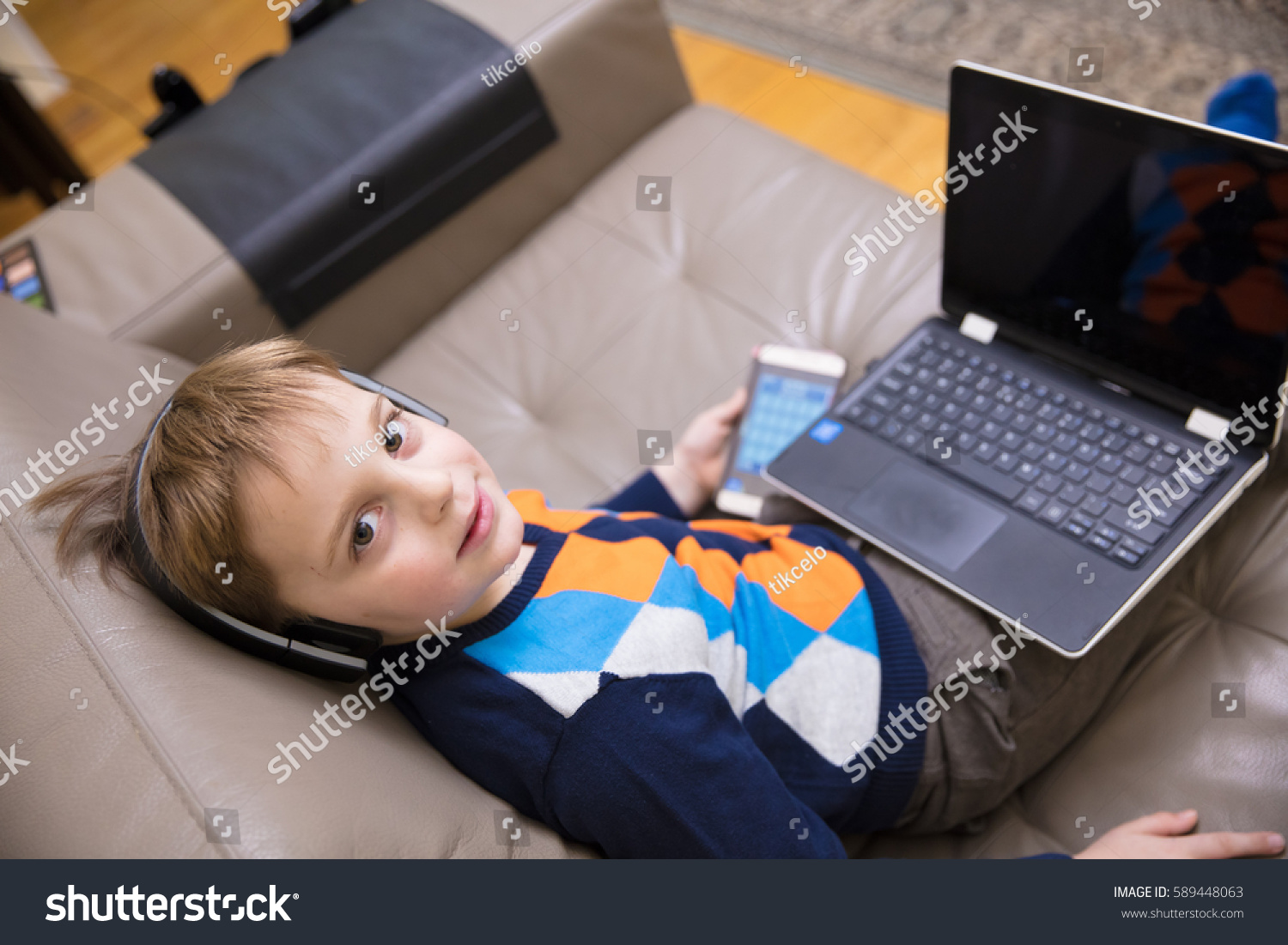 Little boy using a white  laptop computer at home along with mobile phone, wearing headphones, playing some games. #589448063