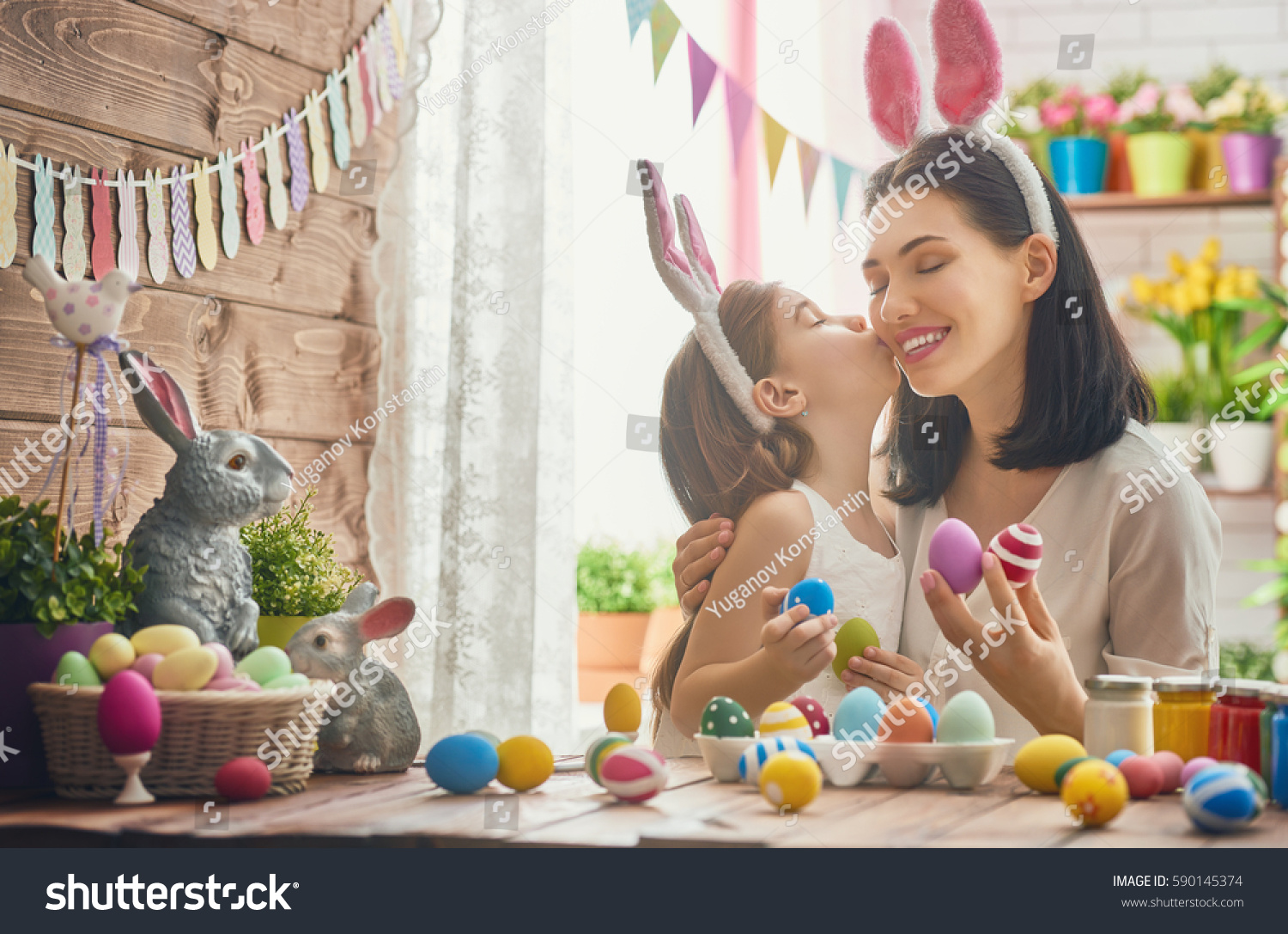 Mother and her daughter painting eggs. Happy family preparing for Easter. Cute little child girl wearing bunny ears. #590145374