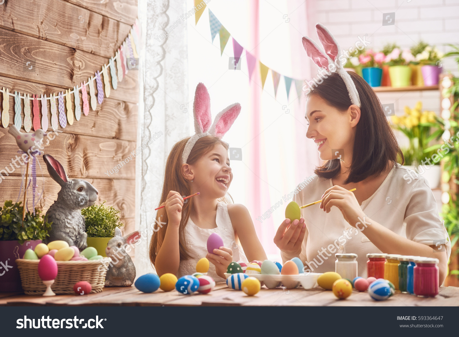 Mother and her daughter painting eggs. Happy family preparing for Easter. Cute little child girl wearing bunny ears. #593364647