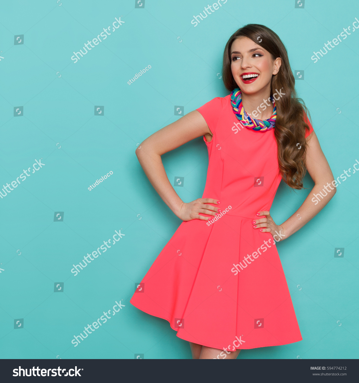 Amused beautiful young woman in pink mini dress posing with hand on hip and looking away. Three quarter length studio shot on turquoise background. #594774212