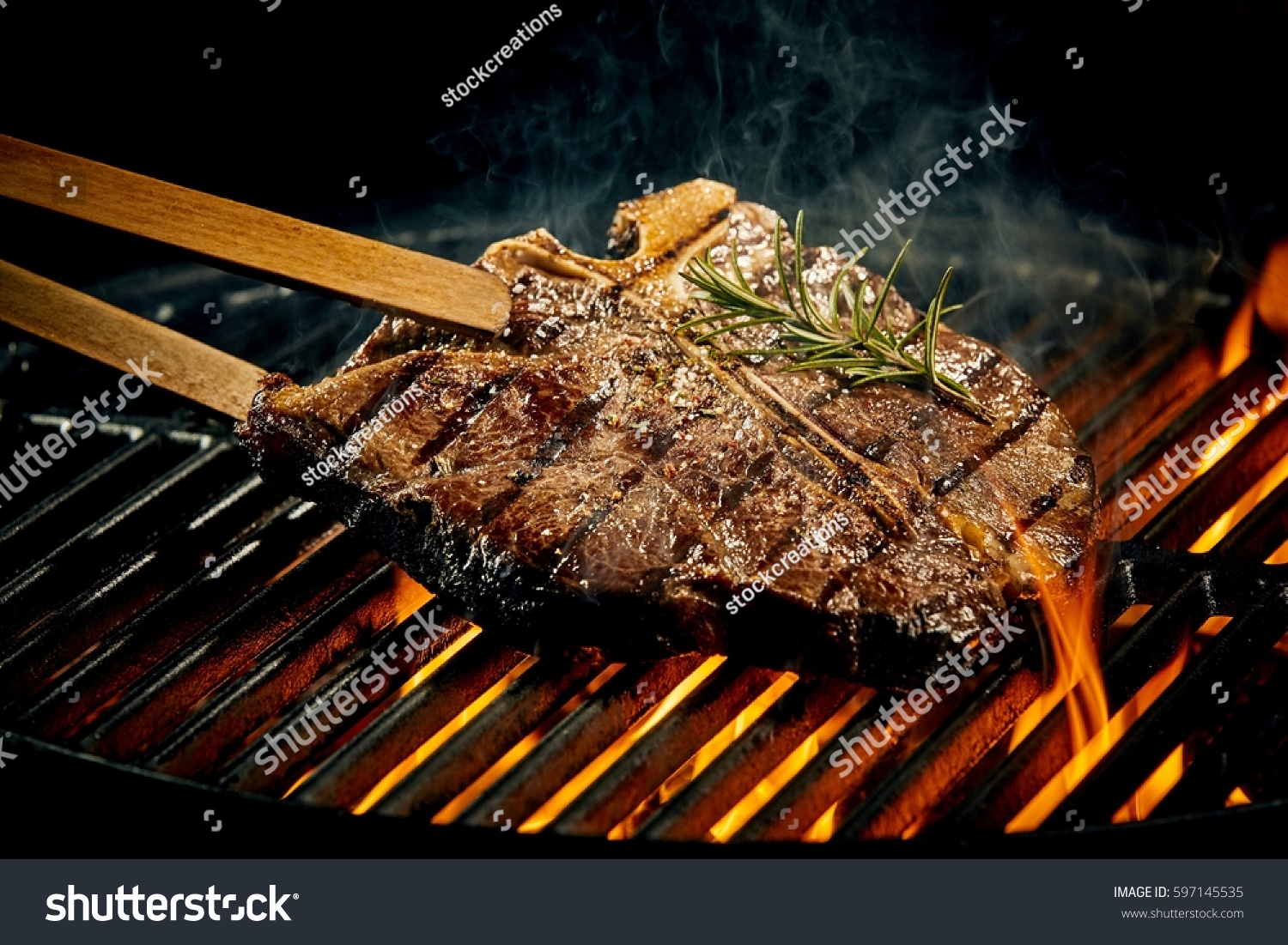 Grilling a tasty tender marinated t-bone steak seasoned with fresh rosemary on a barbecue fire with hot fiery coals in a close up view #597145535