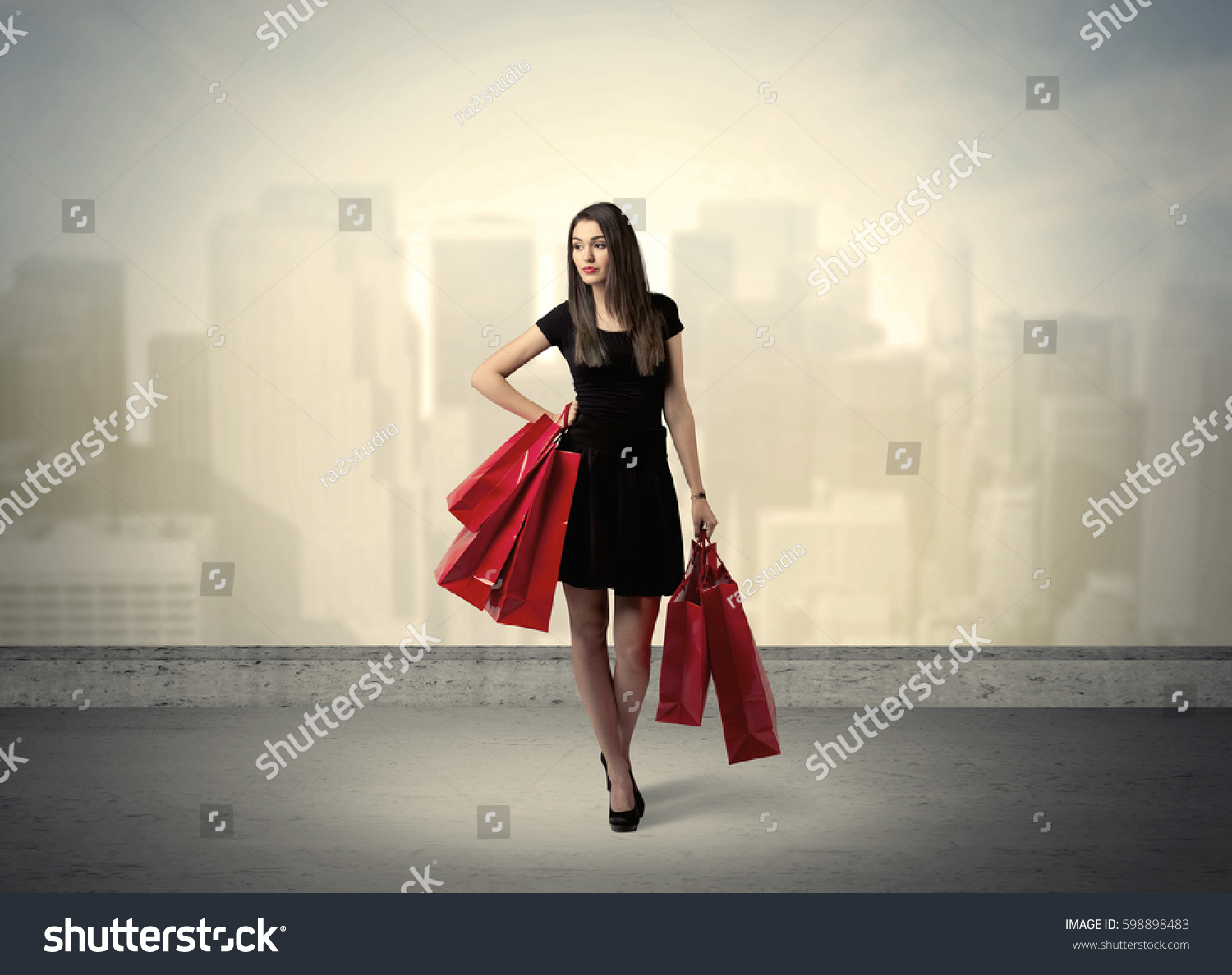 Attractive lady in black holding red shopping bags standing in front o urban landscape with tall buildings concept #598898483