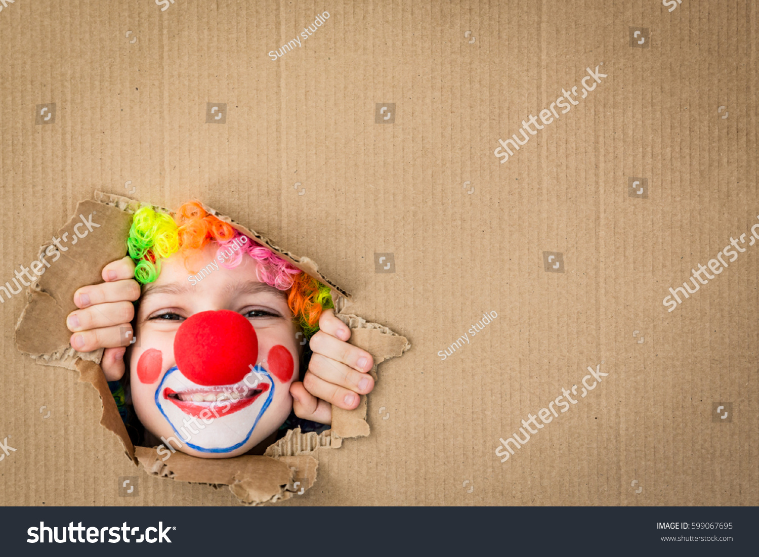 Funny kid clown looking through hole on cardboard. Child playing at home. 1 April Fool's day concept. Copy space. #599067695