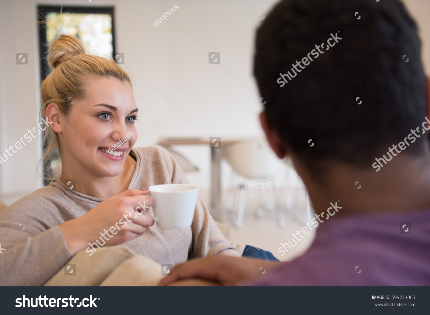Young romantic multiethnic couple sitting on sofa in front of fireplace at home, looking at each other, talking and drinking coffee #599724005