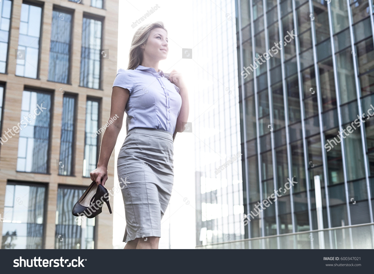 Low angle view of confident businesswoman holding high heels while standing outside office buildings #600347021