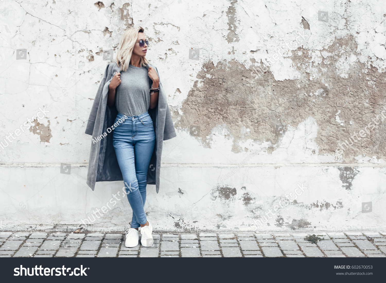 Hipster girl wearing blank gray t-shirt, jeans and coat posing against rough street wall, minimalist urban clothing style, mockup for tshirt print store #602670053
