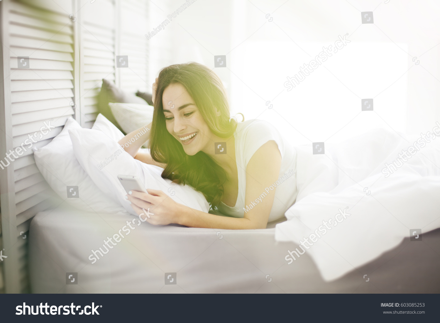 Happy brunette girl using a mobile phone lying on the white bed at home with a window in the background. #603085253