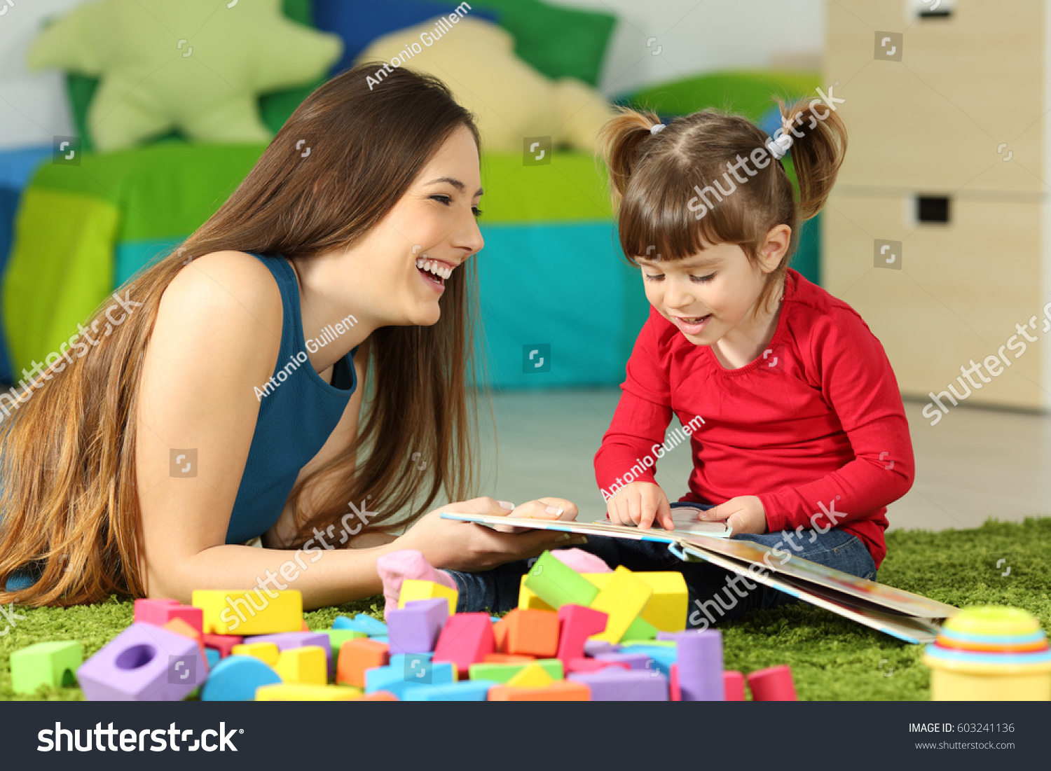 Mother and toddler playing together with a book lying on the floor in the bedroom at home with a colorful background #603241136