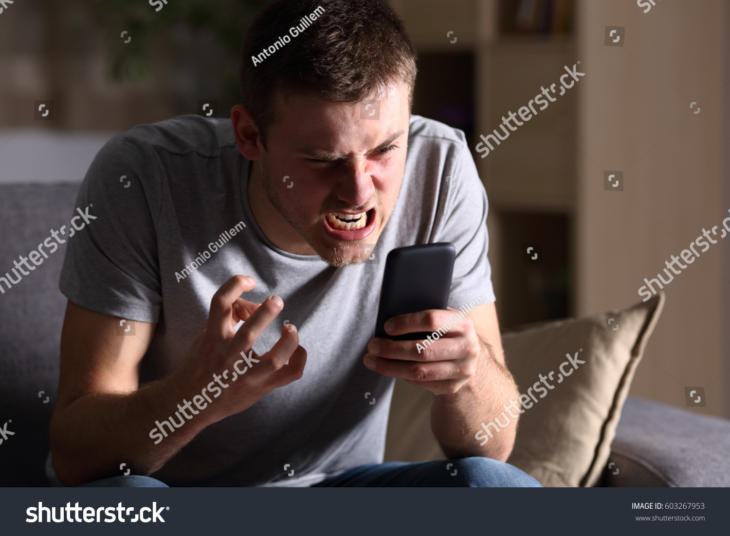 Single angry person with a mobile phone sitting on a sofa in the living room in a house indoor with a dark background #603267953