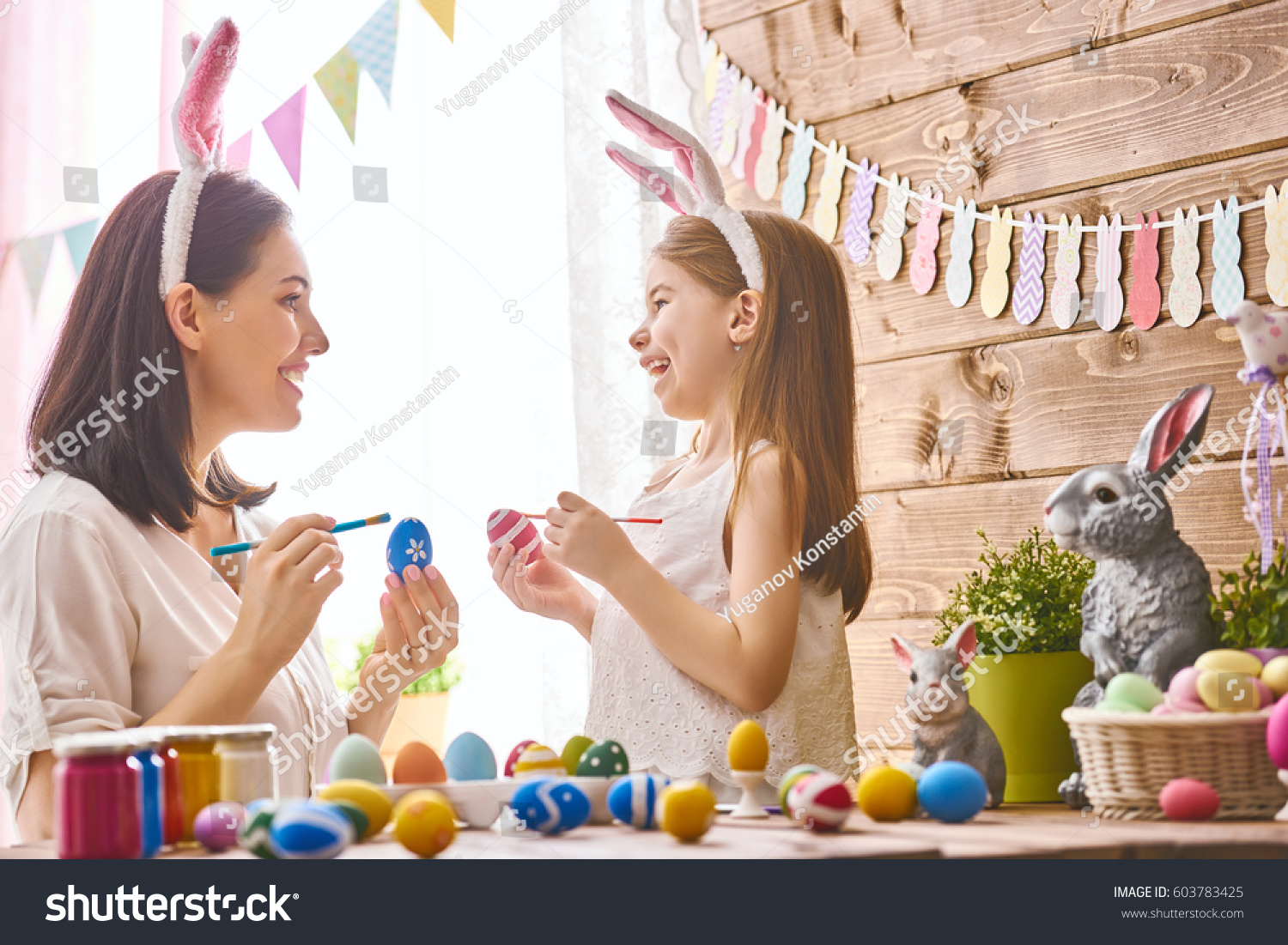 Mother and her daughter painting eggs. Happy family preparing for Easter. Cute little child girl wearing bunny ears. #603783425