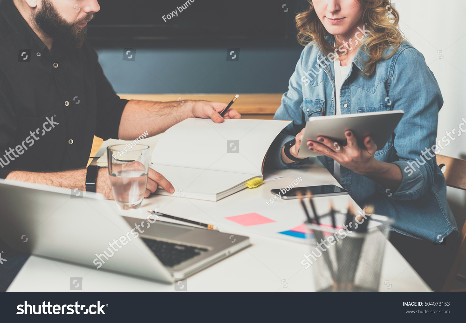 Meet one-on-one. Business meeting. Teamwork. Businessman and businesswoman sitting at table. Woman uses a Tablet PC,man looking catalog.On table is laptop, smartphone,stationery. Freelancers working. #604073153