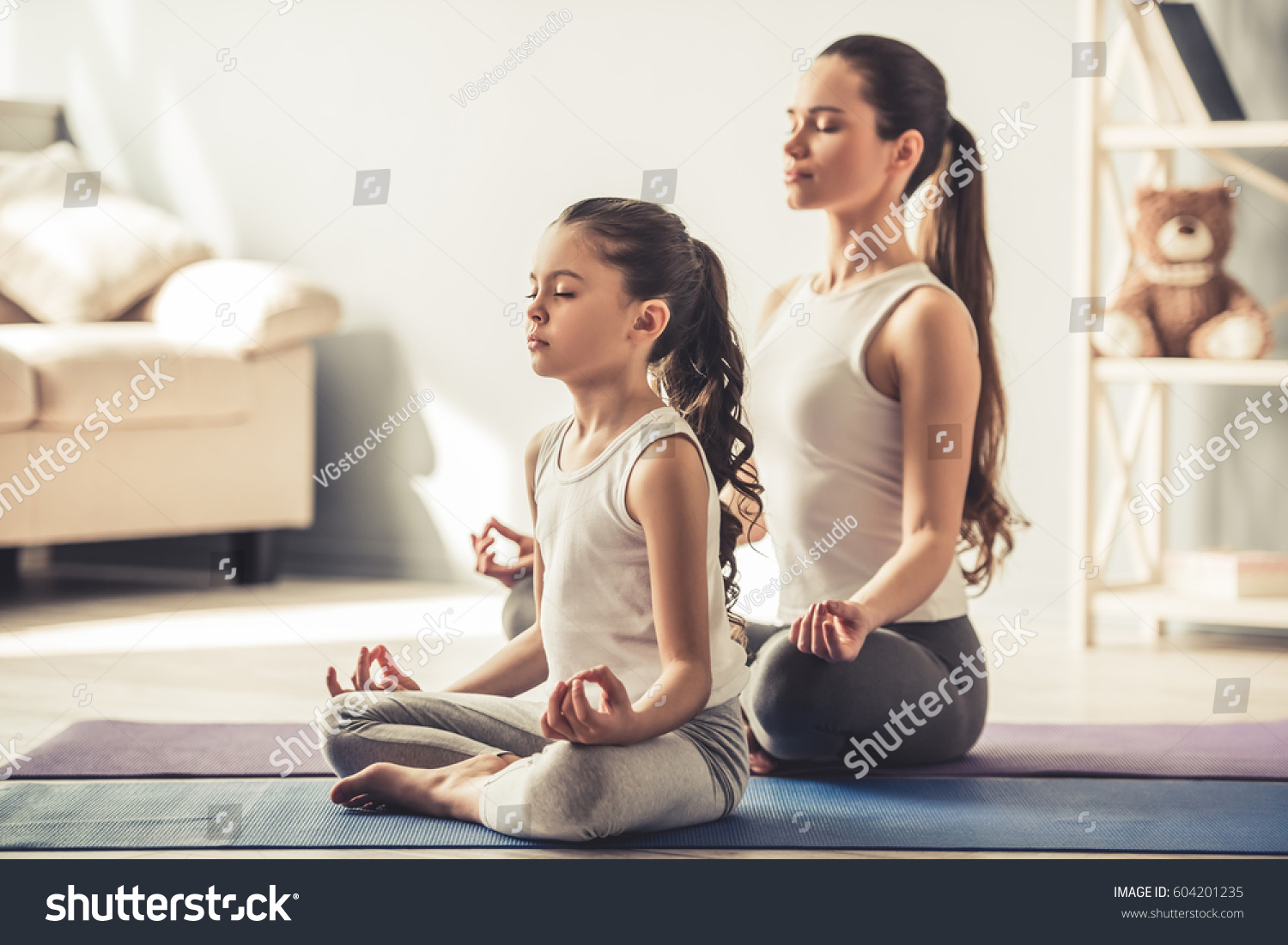 Beautiful young woman and her charming little daughter are smiling while doing yoga together at home #604201235
